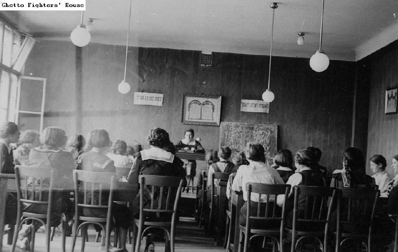 b&w photo of classroom with view of students backs and a teacher at the front of the room