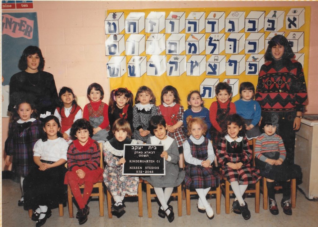 two rows of young girls sitting, flanked by two standing women teachers. Behind them on the wall is the Hebrew alphabet. Two girls in the front row hold a movable-type sign identifying the class.
