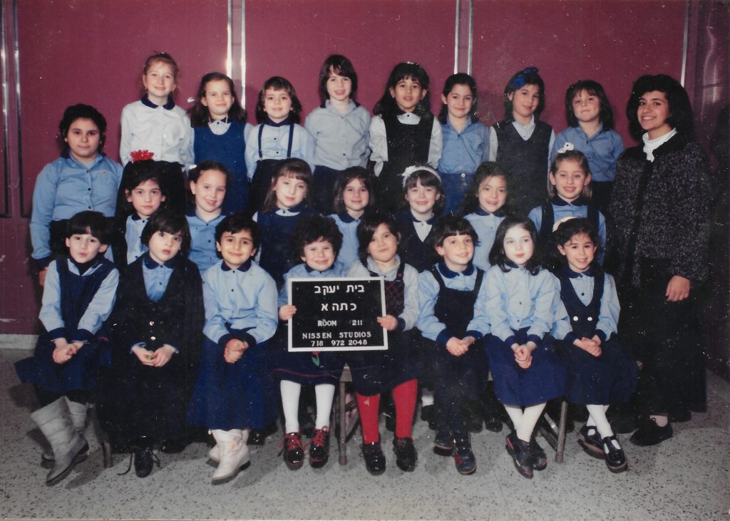Three rows of girls wearing light blue shirts and dark blue skirts or jumpers. A woman teacher stands to the right. Two girls in the front row hold a sign identifying the class.