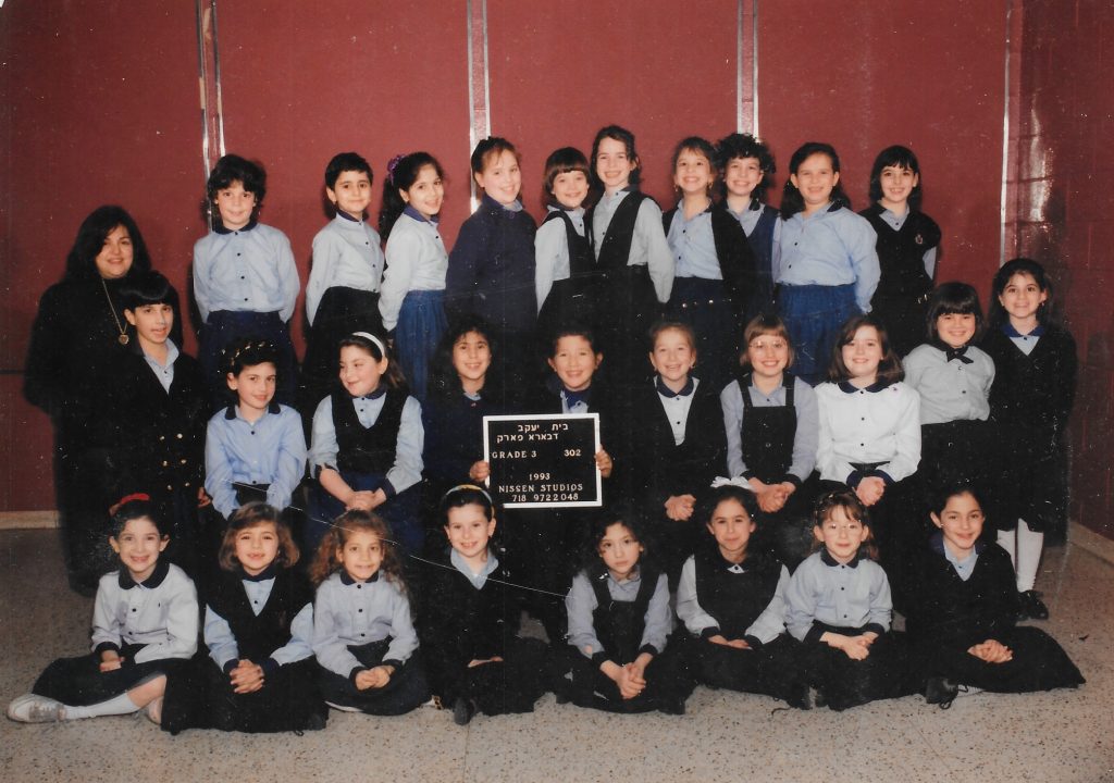 Three rows of girls, one standing on a bench, one sitting on a bench, and one sitting on the floor. The girls wear uniforms of light blue shirts with dark blue collars and dark blue skirts or jumpers. A teacher stands to the left. Two girls in the middle row hold a black-and-white sign identifying the class.