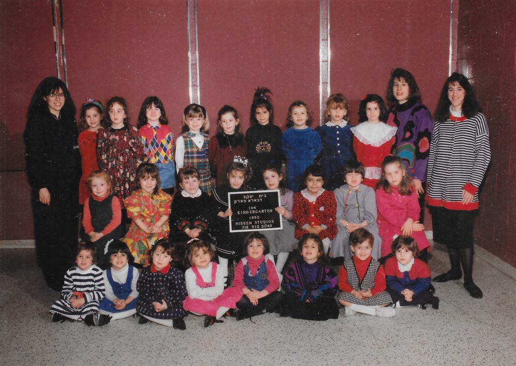 Three rows of girls, one standing on a bench, one sitting on a bench, one sitting on the floor. Two girls in the middle row hold a black-and-white sign identifying the class. Two teachers flank the class on either side.