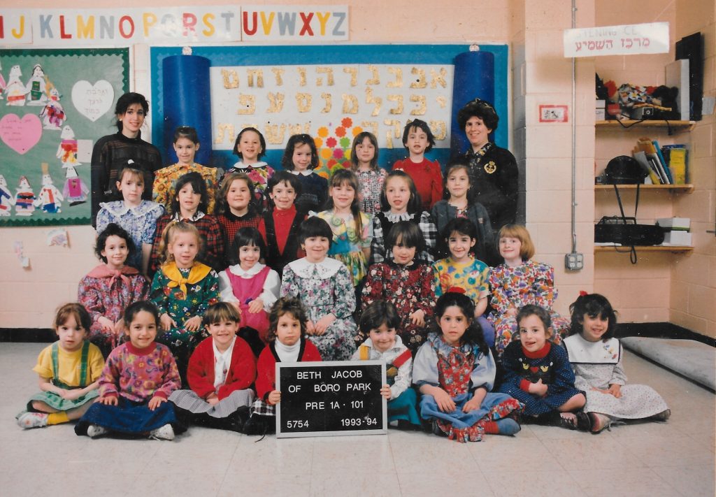 Four rows of girls, one standing on a bench, one standing on the floor, one sitting on a bench, and one sitting on the floor. Two girls in the front row hold a black-and-white sign identifying the class. Two teachers flank the class on either side of the back row.